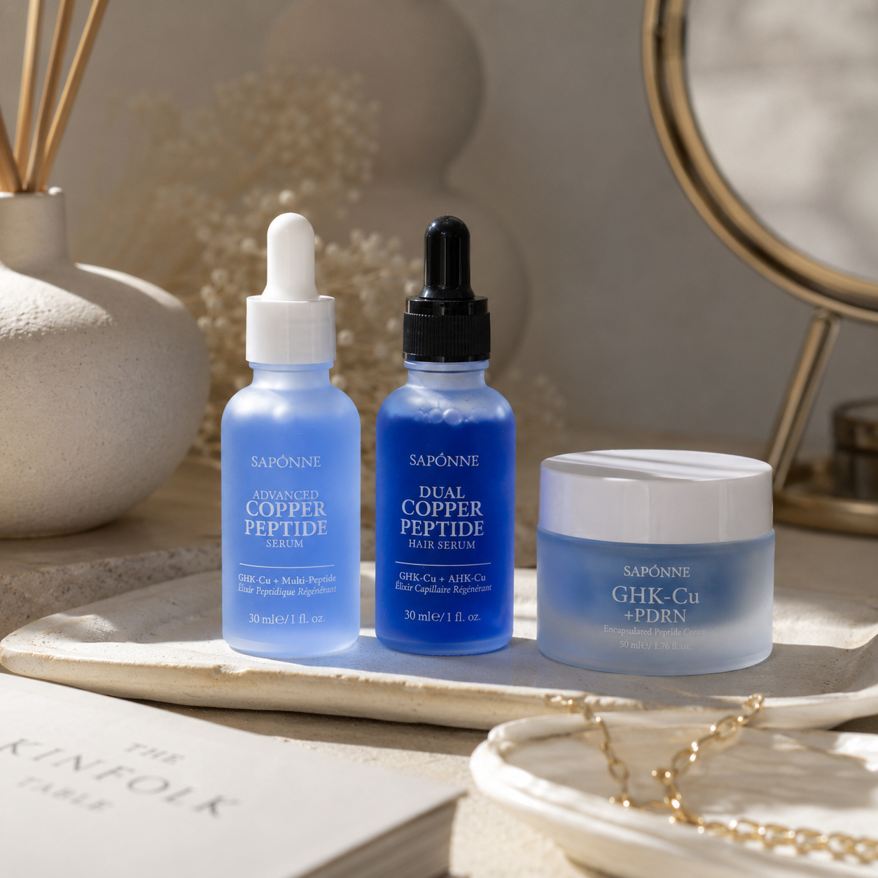 Three Sapónne skincare products arranged on a light stone tray: a blue serum dropper bottle, a blue hair serum dropper bottle, and a frosted blue jar with a white lid. The scene is styled with a mirror, gold jewelry, and soft neutral decor in the background.