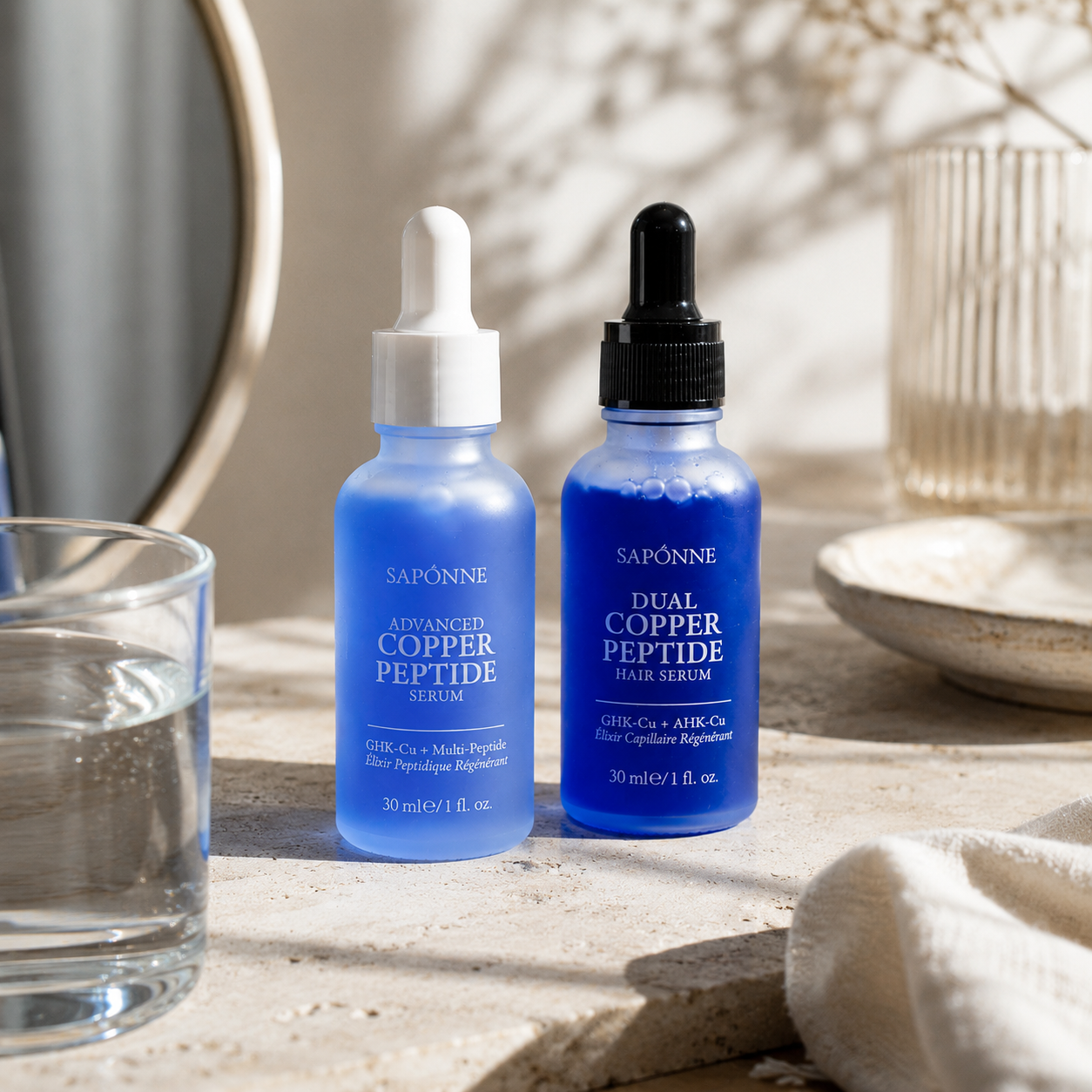 Two blue Saponne skincare dropper bottles on a stone surface with a glass of water, a mirror, and soft natural shadows in the background. The bottles are labeled Advanced Copper Peptide Serum and Dual Copper Peptide Serum.
